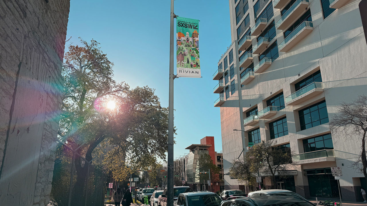 Sonnige Straßenszene in einer Stadt mit parkenden Autos, Bäumen und modernen Gebäuden. An einem Laternenmast hängt ein Banner für das SXSW-Festival.