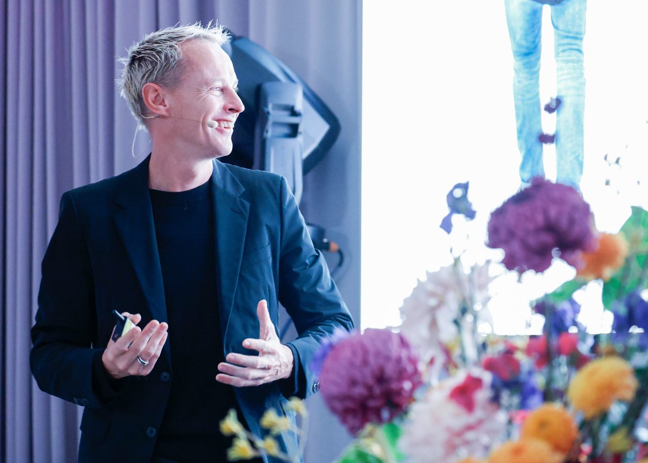 Daan Roosegaarde: Porträt eines Mannes mit kurzem, blonden Haar und einem freundlichen Lächeln. Er trägt ein dunkles Sakko über einem schwarzen Hemd. Im Vordergrund rechts sind verschwommene Blumen zu sehen. Im Hintergrund hängt ein grauer Vorhang. Der Mann präsentiert mit einem kleinen Mikrofon, das an seiner Wange befestigt ist.