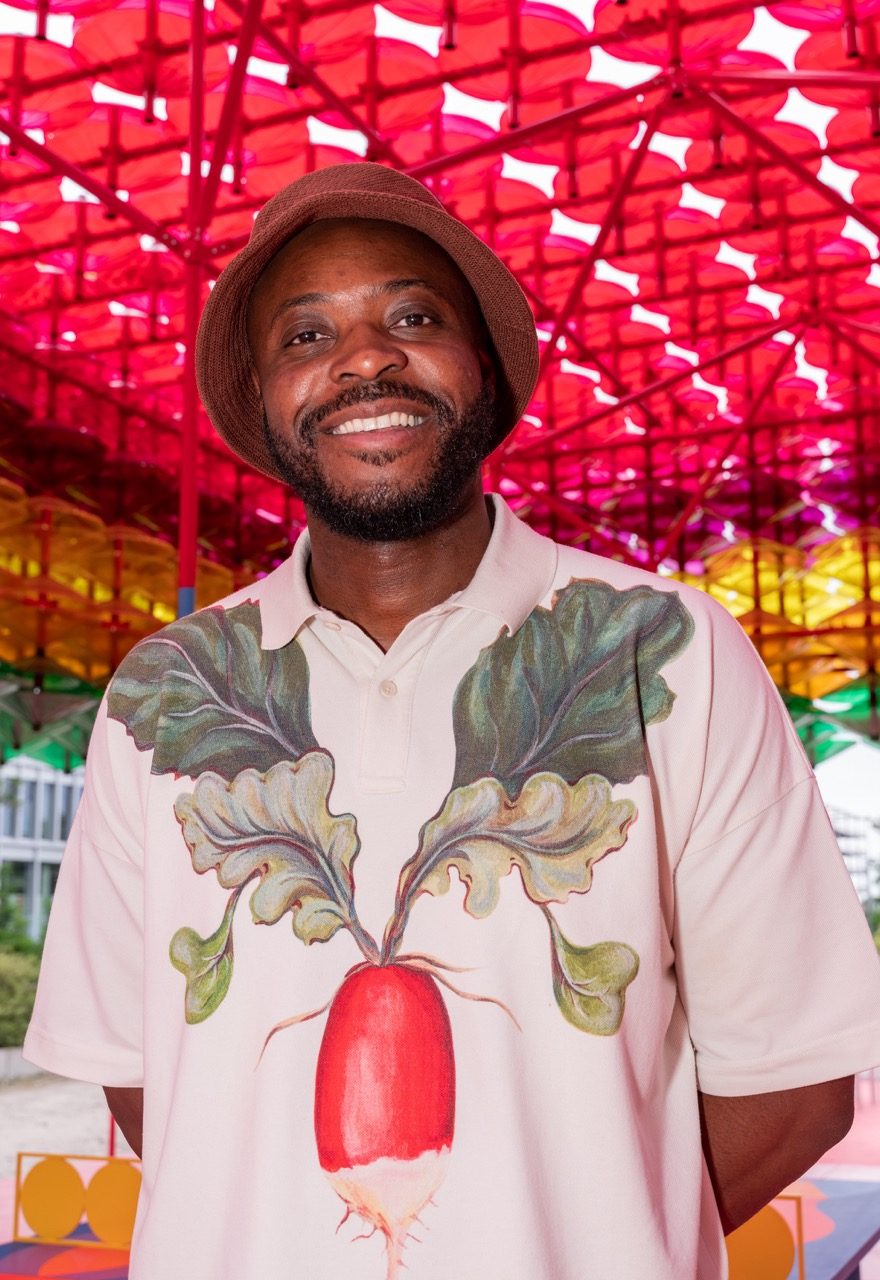 Yinka Ilori: Porträt eines Mannes, der in die Kamera lächelt. Er trägt ein helles Hemd mit einer riesigen Radieschen-Aufschrift in der Mitte und einen braunen Fischerhut. Der Hintergrund ist bunt, mit einer Decke, die hauptsächlich rot, aber auch gelb und grün ist.
