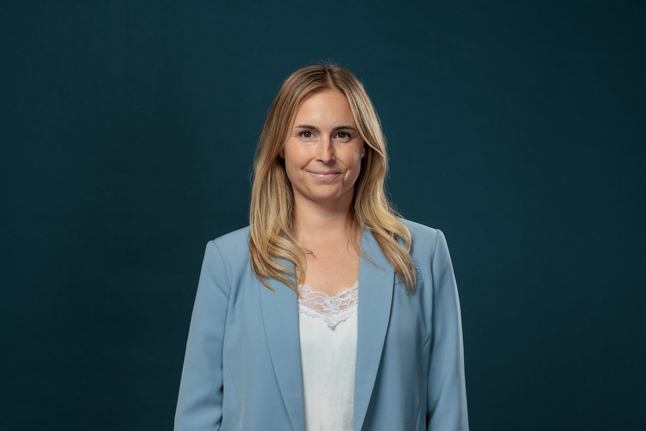 Porträt einer Frau mit schulterlangen blonden Haaren und einem dezenten Lächeln. Sie trägt einen hellblauen Blazer über einer weißen Bluse mit Spitzenbesatz. Der Hintergrund ist dunkelgrün und einfarbig.