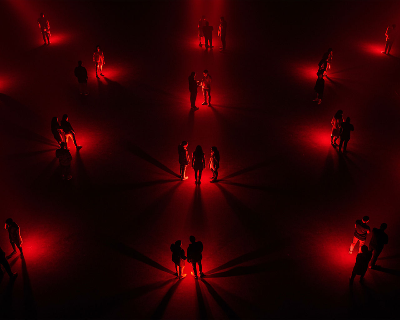 multiple small groups of people standing together in red spotlights in a dark room chatting