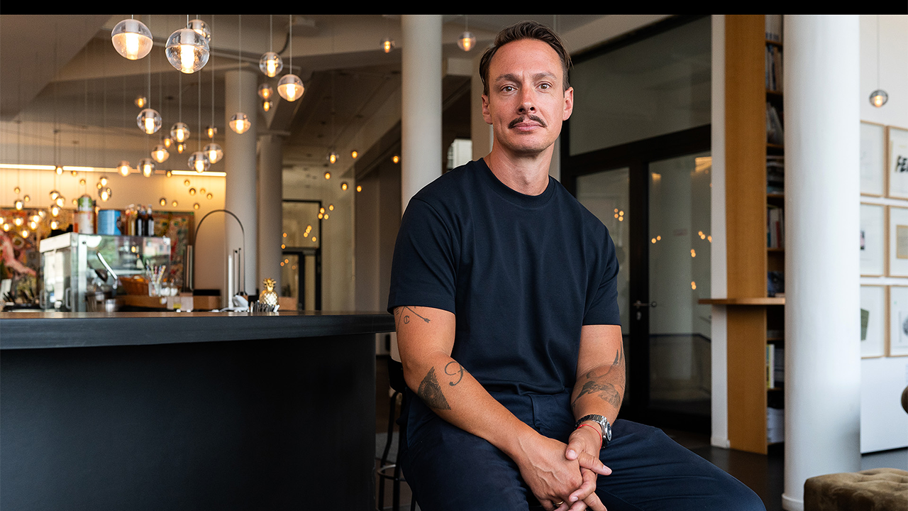 Person sitzt auf einem Hocker in einem modernen Innenraum mit Theke und PendelleuchA person is sitting on a stool in a modern interior, with a counter and pendant lights in the background. The person is wearing a dark T-shirt and dark trousers, with their hands clasped in their lap.