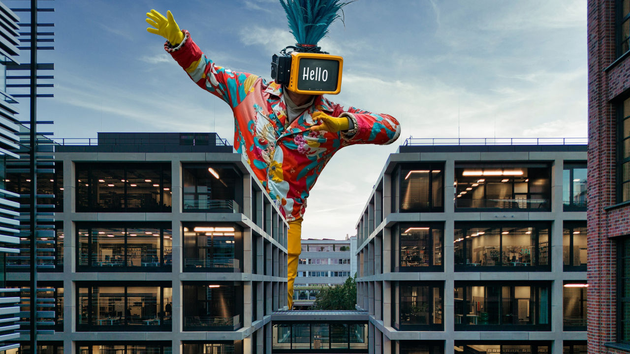 A huge figure with a TV for a head saying ‘hello’ while standing between the offices of the ‘House of Communication’