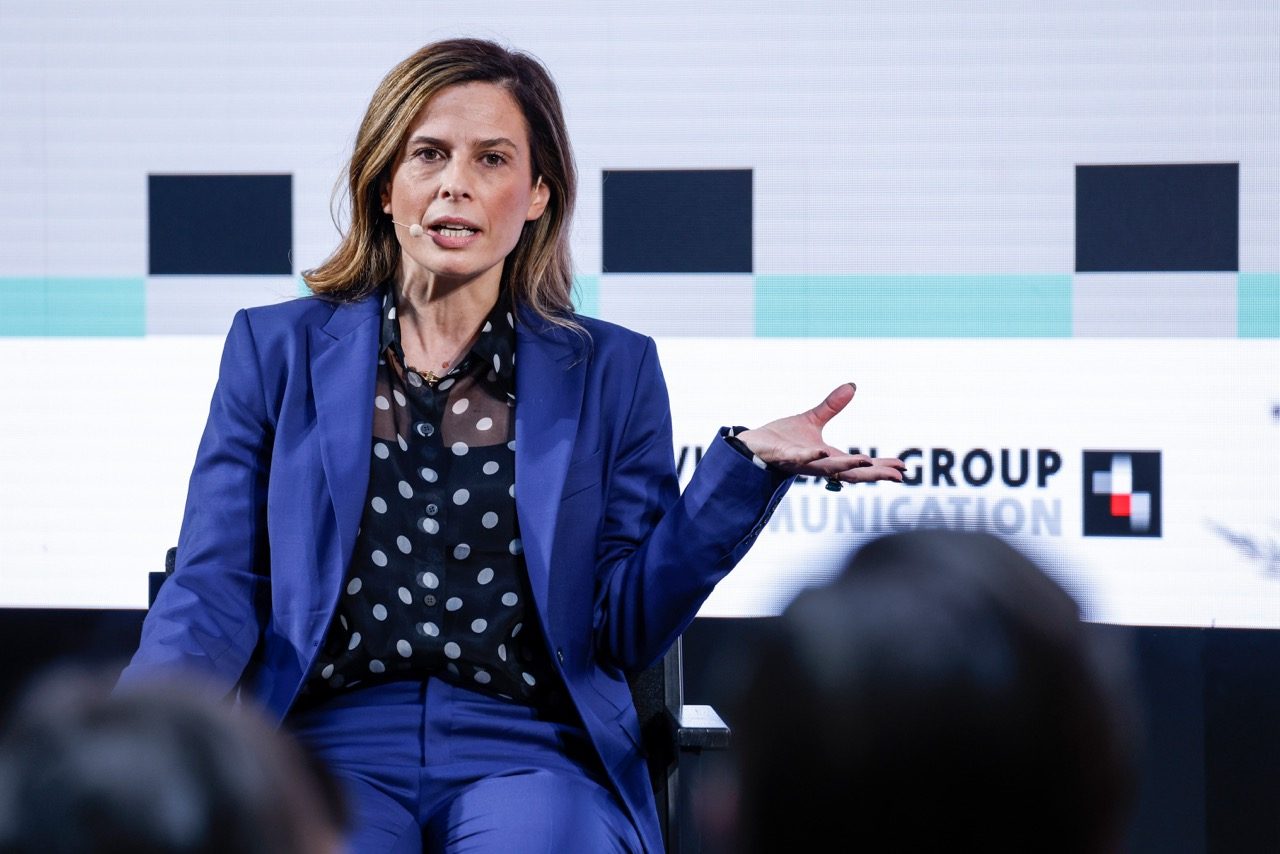 Francesca Bria: A woman with brunette hair and blonde highlights is talking on a stage in a blue suit with a polka dot chemise underneath.