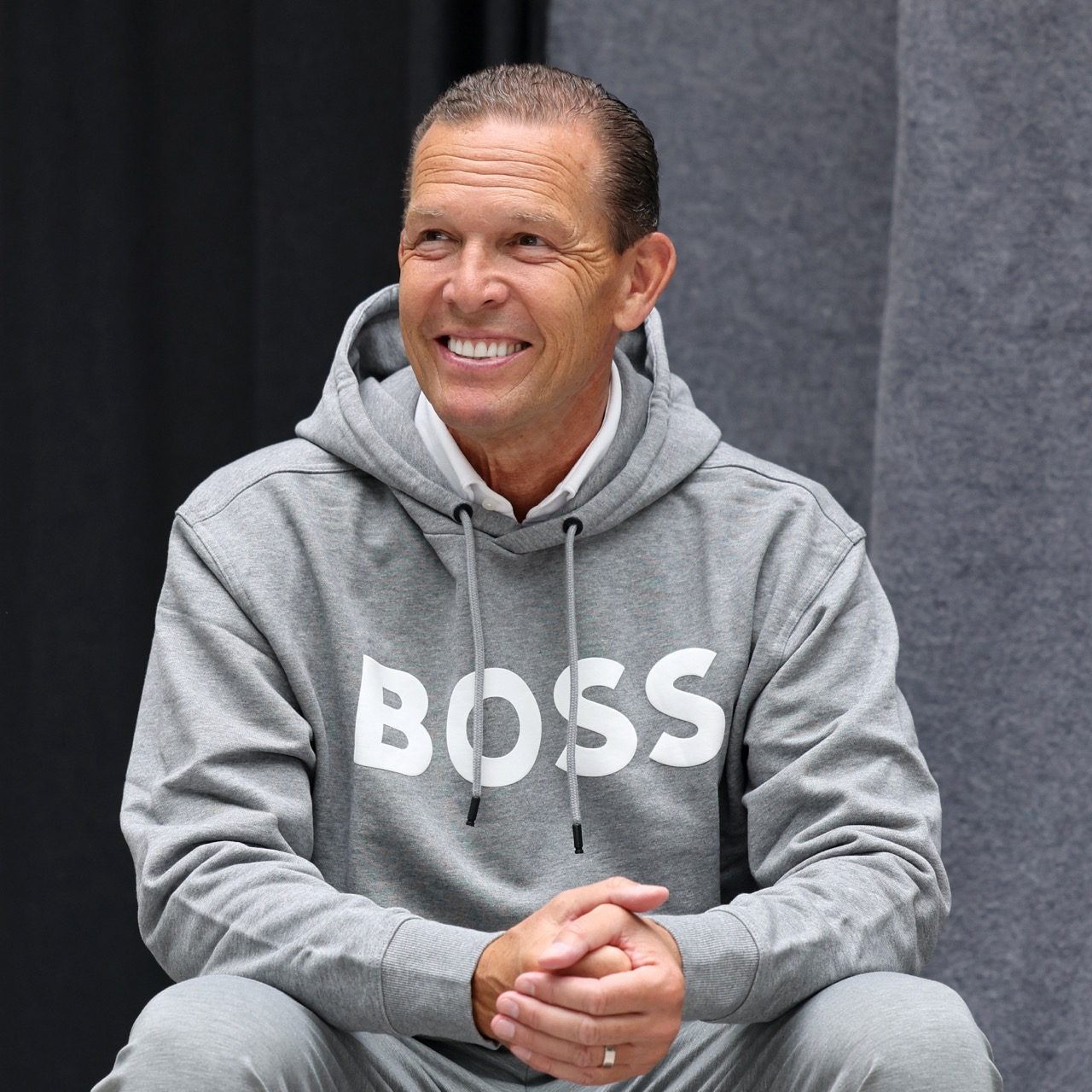Daniel Grieder: Portrait of a man with very short hair, who is looking to the left of the camera and smiling in the distance. He is sitting in a grey hoodie with the logo of the brand "Hugo Boss".