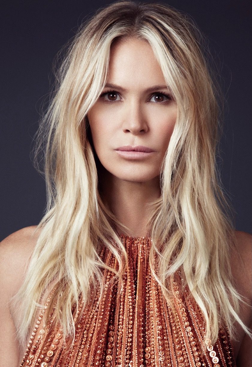 Portrait of a woman with long blonde hair, she looks directly into the camera. She is wearing an orange dress with no sleeves.