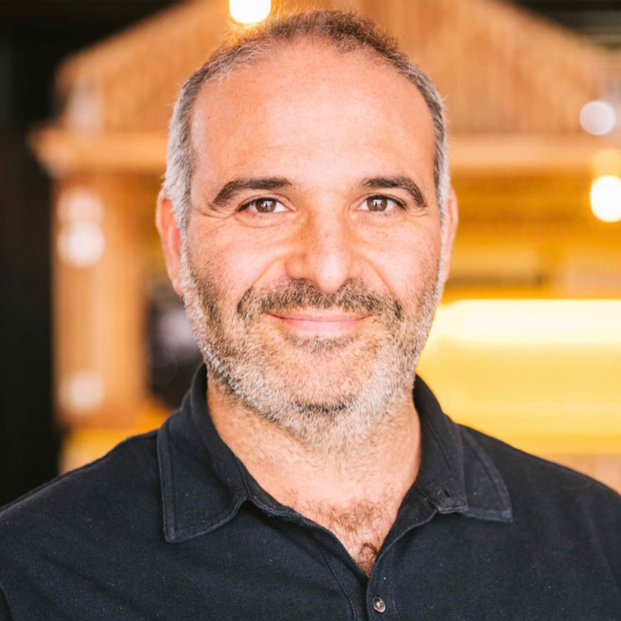 Philippe Fass. Portrait of a man with salt-and-pepper hair and beard, smiling in a warmly lit environment. He is wearing a black polo shirt.