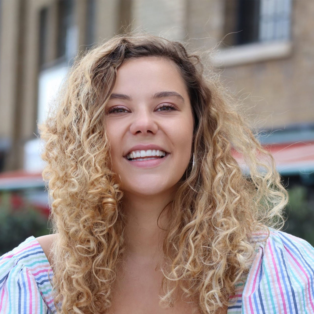 Elli Britton. Close-up of a smiling woman with curly blonde hair in front of a blurred urban background. She wears a colorful striped blouse with puffed sleeves.