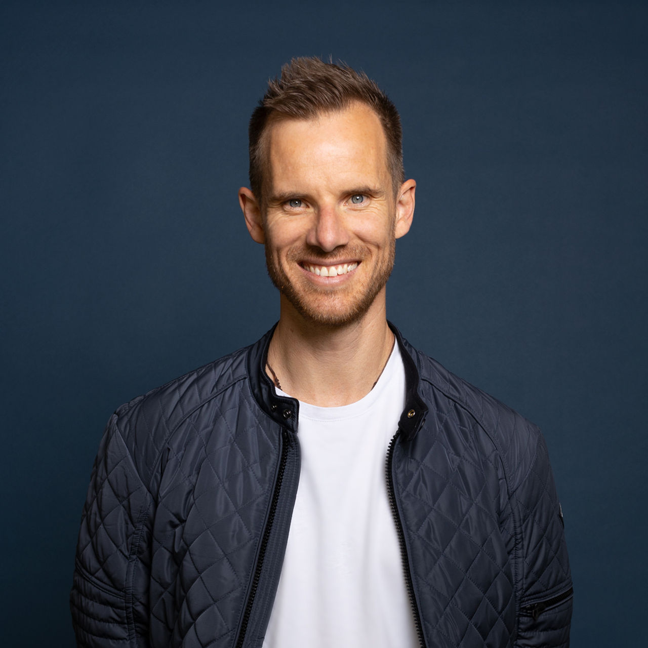 Andy Wyeth. Portrait of a smiling man with short hair standing in front of a dark blue background. He wears a white T-shirt and a dark blue quilted jacket.