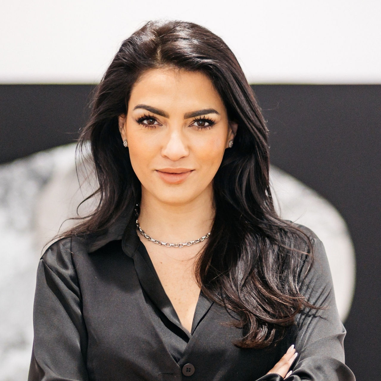 Ruth Santana. Woman with long dark hair stands with crossed arms in front of a modern black-and-white artwork. She wears a black shirt and a silver necklace.